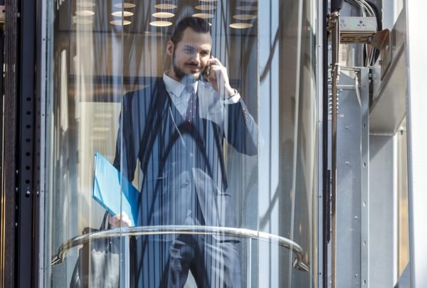 Businessman in modern glass elevator talking by the phone and holding papers